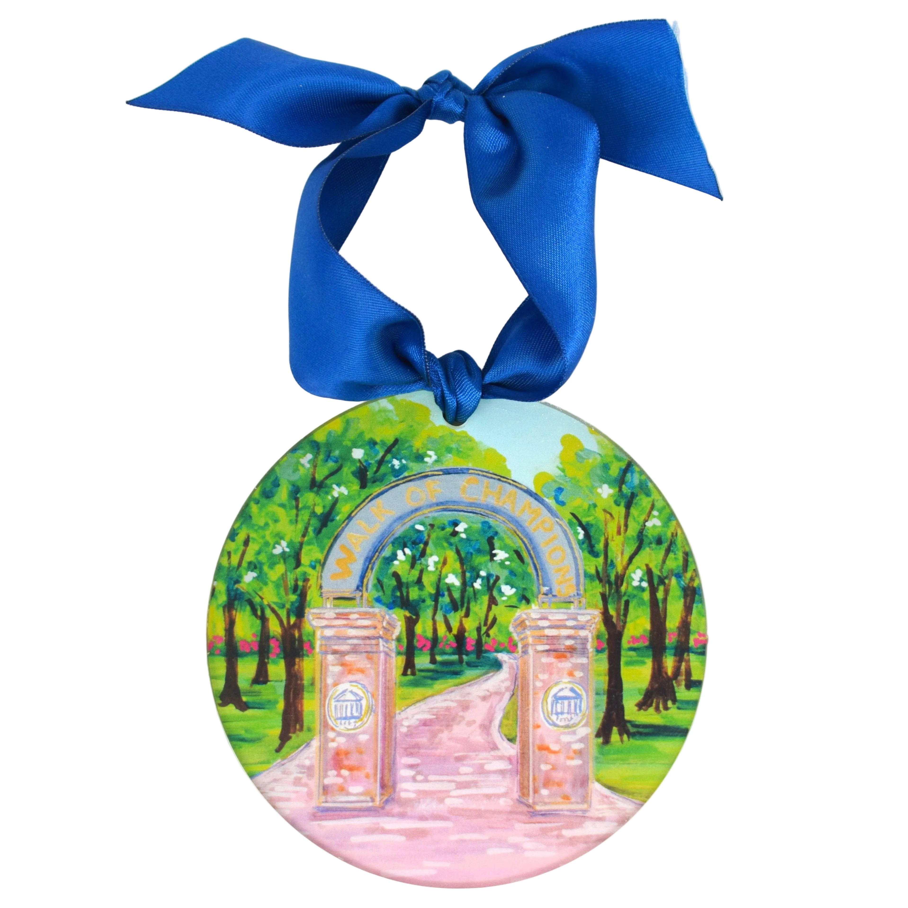 A round decorative ornament featuring a painted scene of a pink pathway leading through a green park under an arched bridge, hanging from a large royal blue satin bow on a white background. University of North Carolina Ornament 