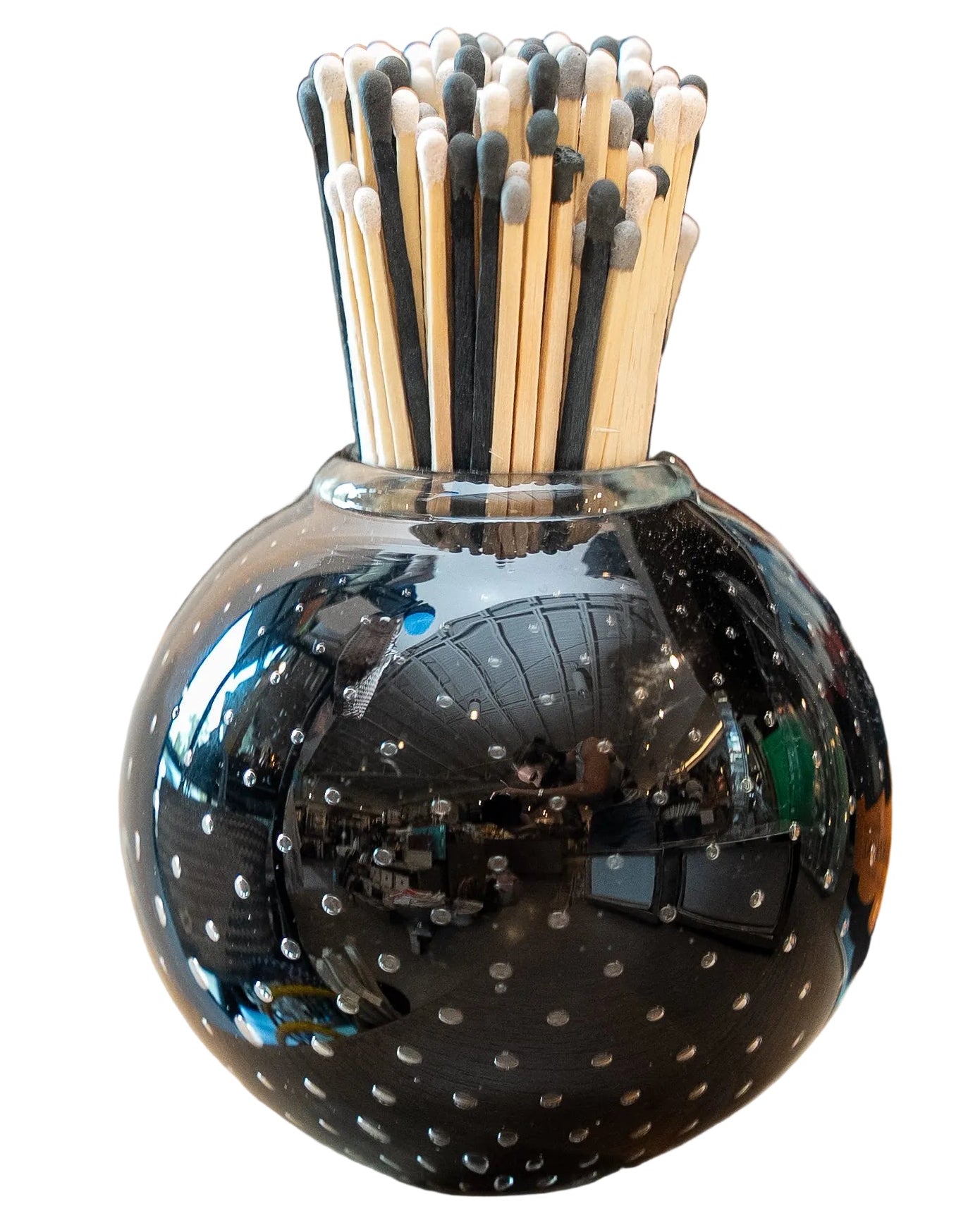 A glossy black glass vase with white speckled dots, filled with wooden matchsticks that have black tips, set on a light wood surface with a blurred background.