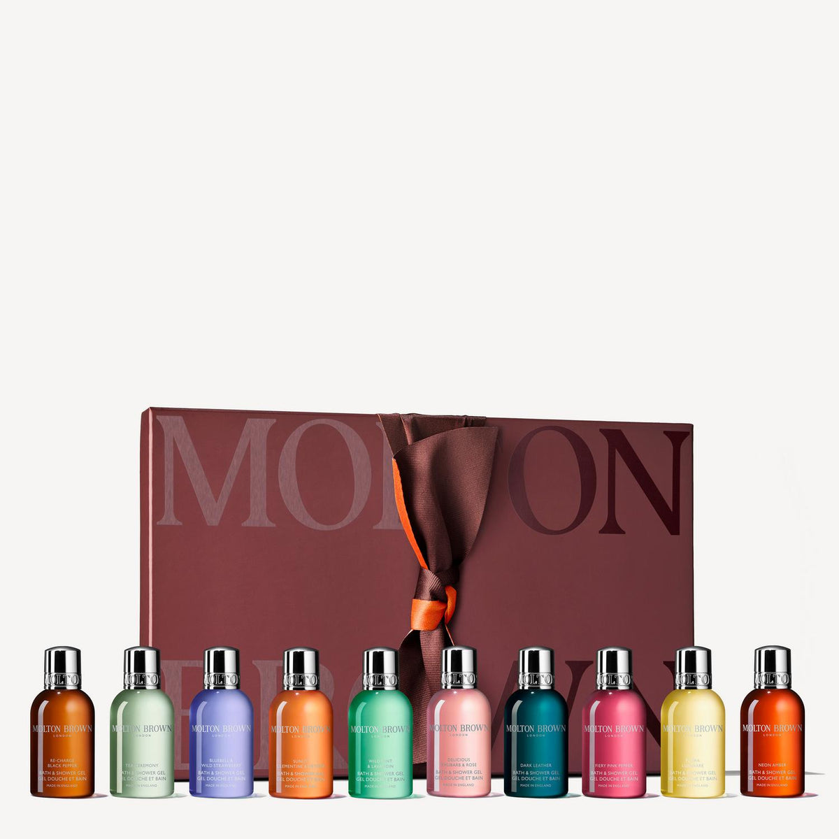 A set of ten small cosmetic bottles in assorted colors lined up in front of a large mauve-brown box with the brand name MON BON visible, tied with a dark ribbon and orange accent.