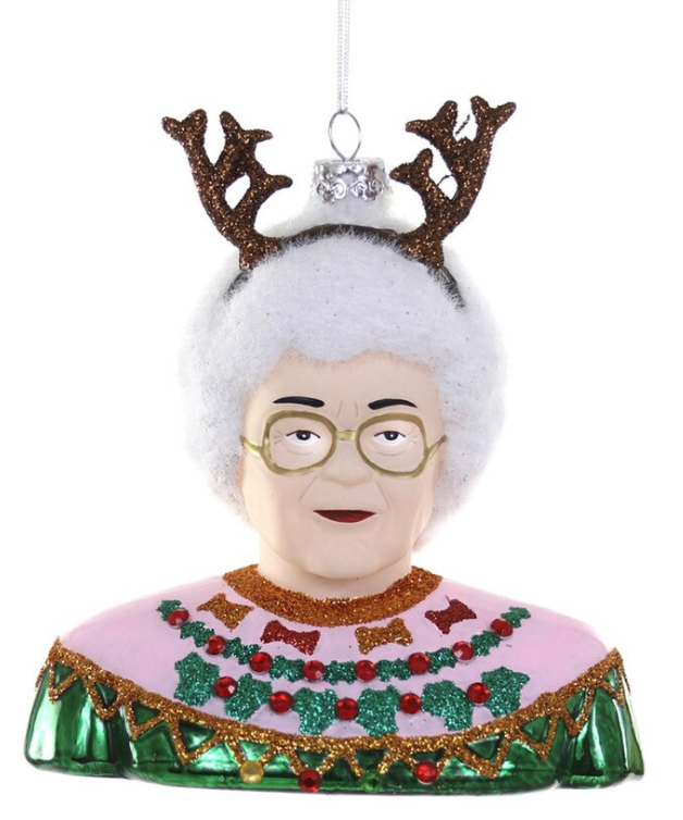 A Christmas ornament shaped like a bust portrait of an elderly woman with white hair, round glasses, and brown glittery antlers, wearing a pink top with green and red holiday patterns and a green skirt, hanging against a white background.