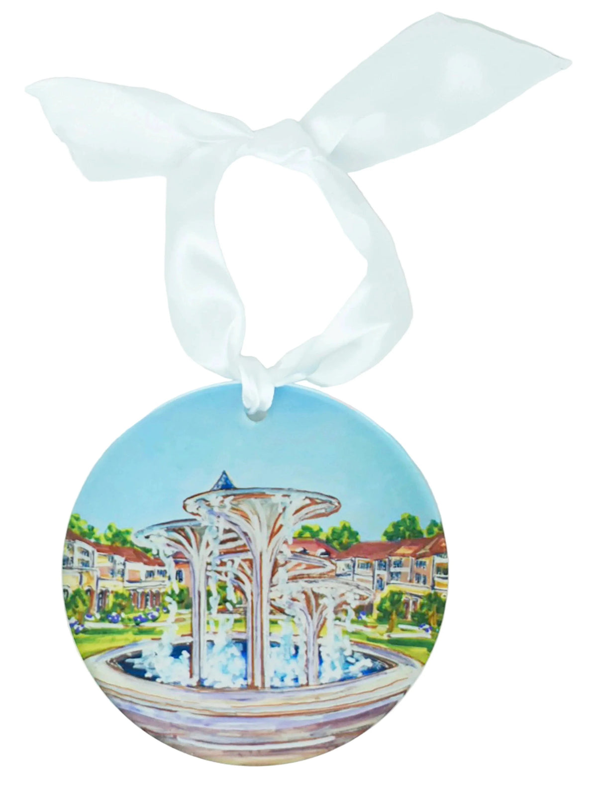 A round ceramic ornament featuring a painted illustration of a fountain in front of a resort-style building, hanging from a white satin ribbon bow on a white background.