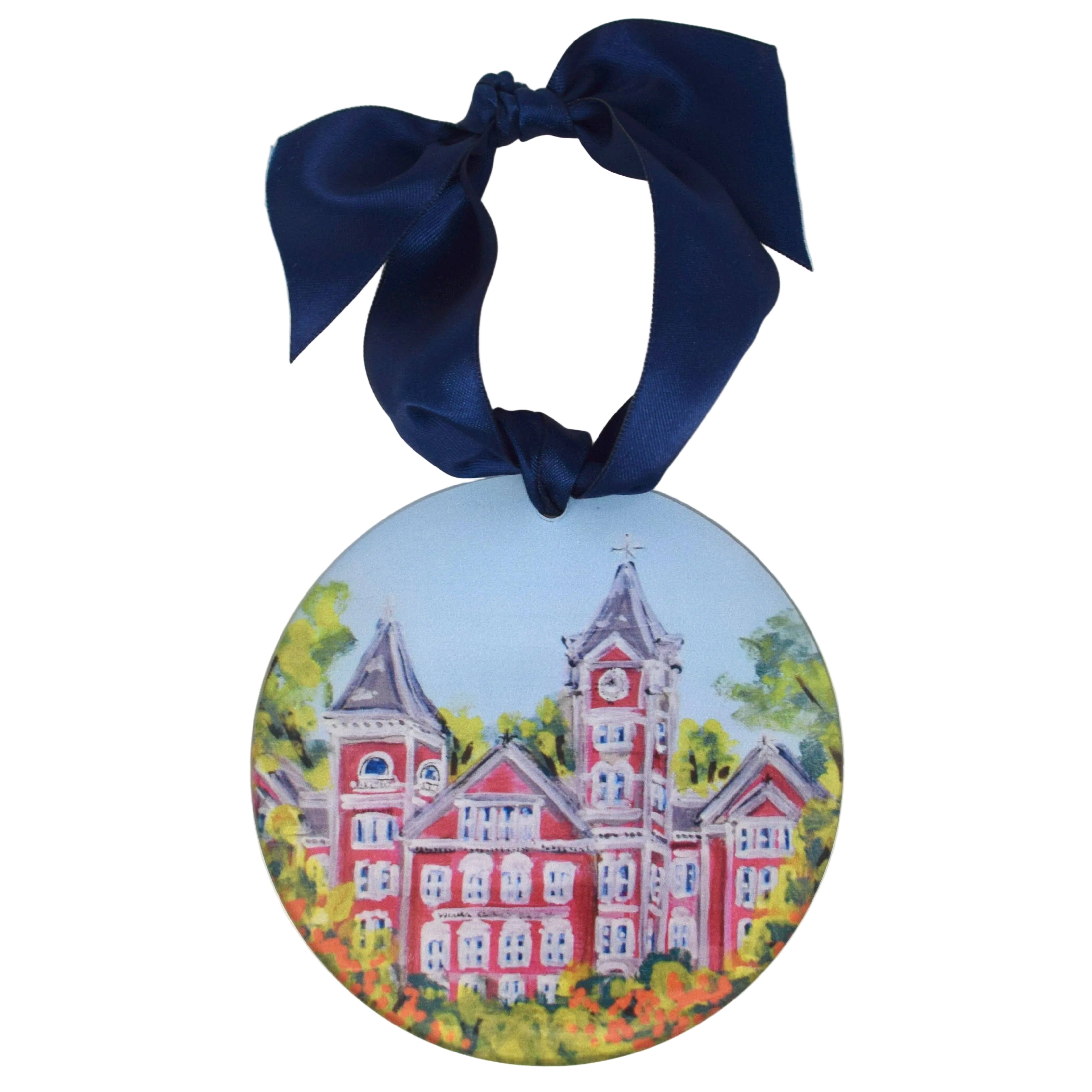 A round ceramic ornament featuring a painted illustration of a red brick schoolhouse with green trees and a blue sky, hanging from a dark navy blue satin ribbon bow on a white background. auburn ornament