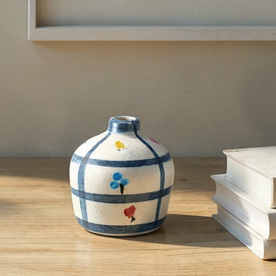 Small decorative vase with colorful patterns on a wooden surface, with books and a framed picture in the background.