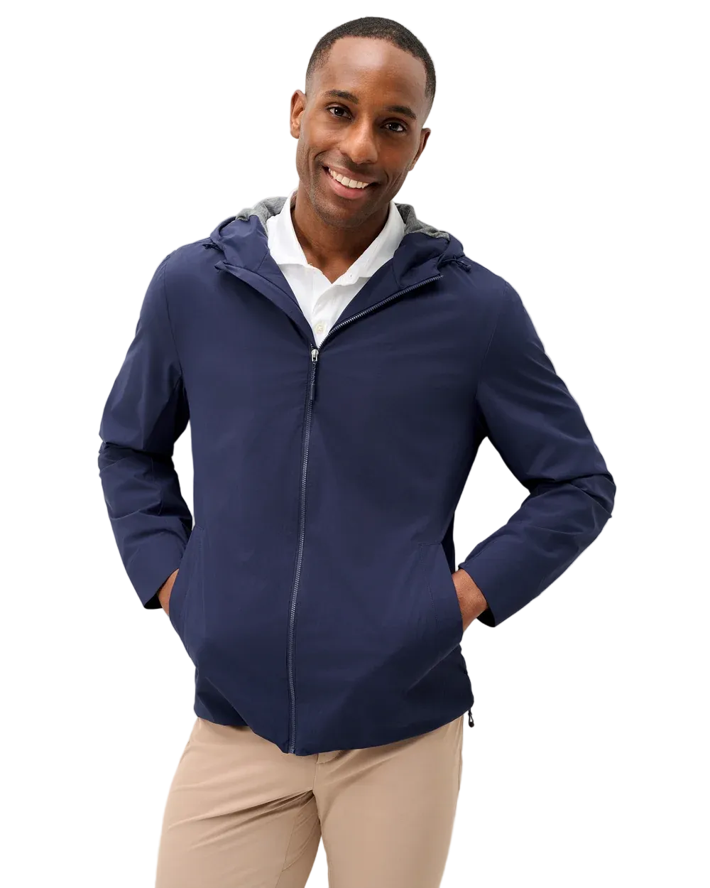A smiling man wearing a navy blue zip-up hooded jacket over a white collared shirt and beige trousers, standing against a light gray studio background.