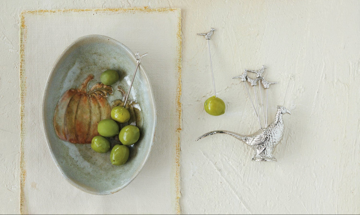 Set of silver pheasant-shaped skewers in a box with a decorative plate and olives on a light surface.