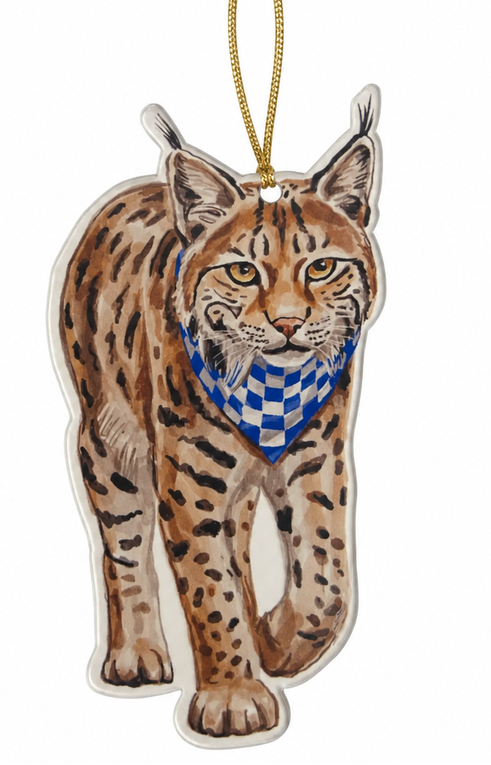 A die-cut hanging ornament shaped like a walking lynx cat, with a gold hanging string at the top. The lynx has tan and brown spotted fur, yellow eyes, and a blue-and-white checkered bandana around its neck, shown on a plain white background.