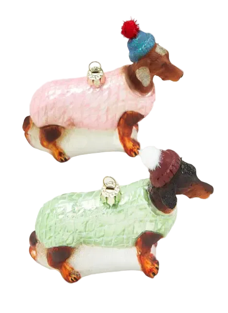 Two dachshund-shaped ornaments dressed in winter sweaters, one in pink with a blue hat and red pom-pom, and one in mint green with a burgundy hat and white pom-pom, on a light gray background.