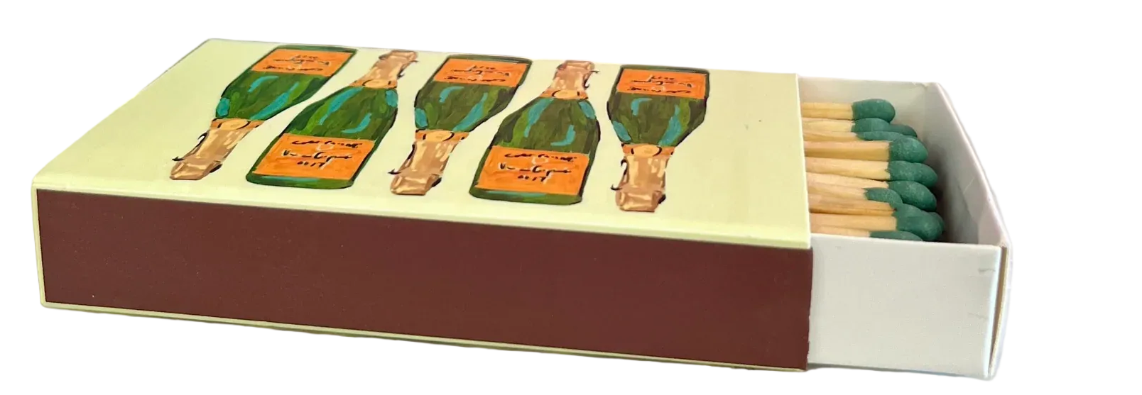 A decorative matchbox with illustrated green bottles and lit green matches sits on a white marble surface in front of a bright pink wall, with a potted green plant in the background.