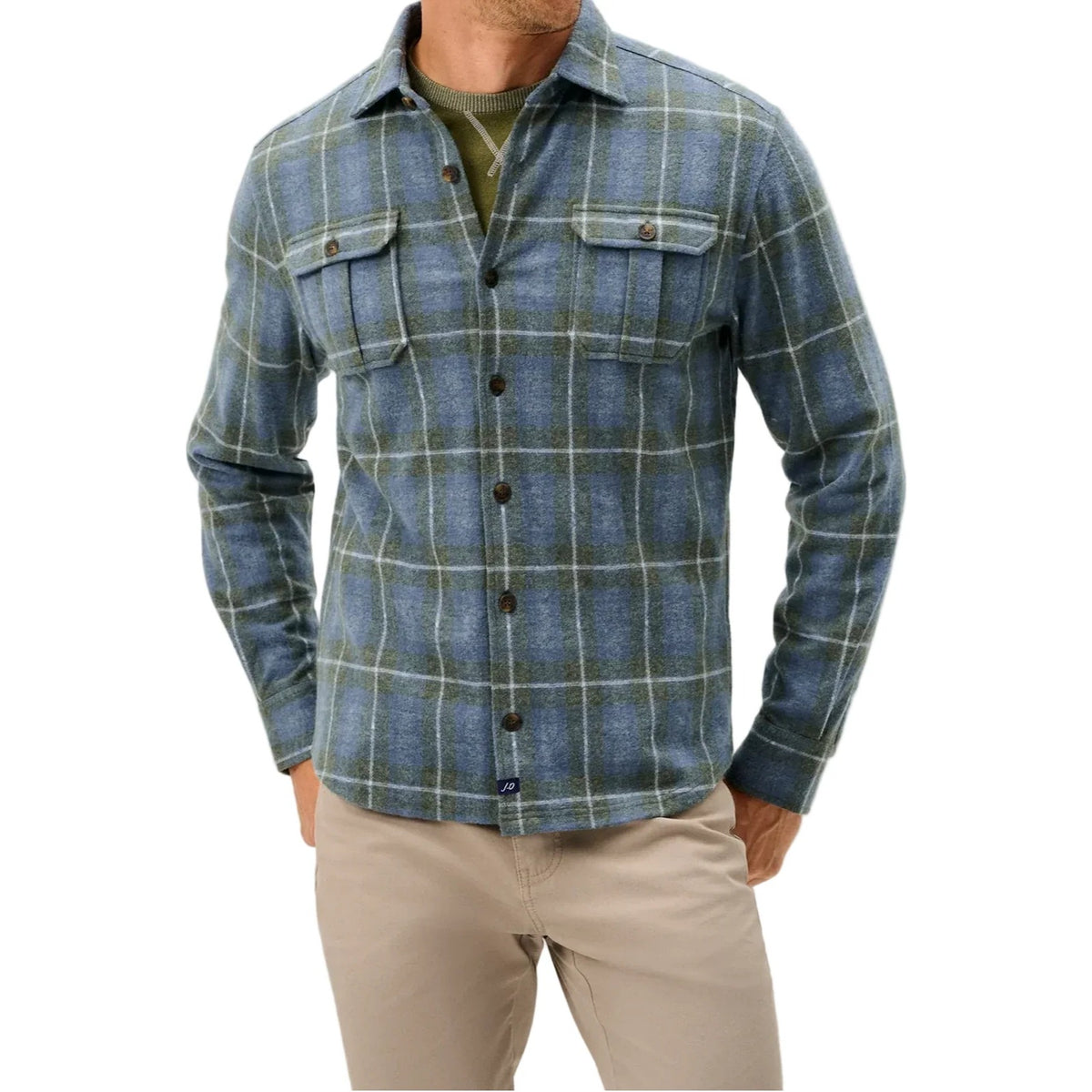 A man wearing a blue and green plaid button-down flannel shirt over an olive green undershirt, paired with beige pants, against a light gray studio background.