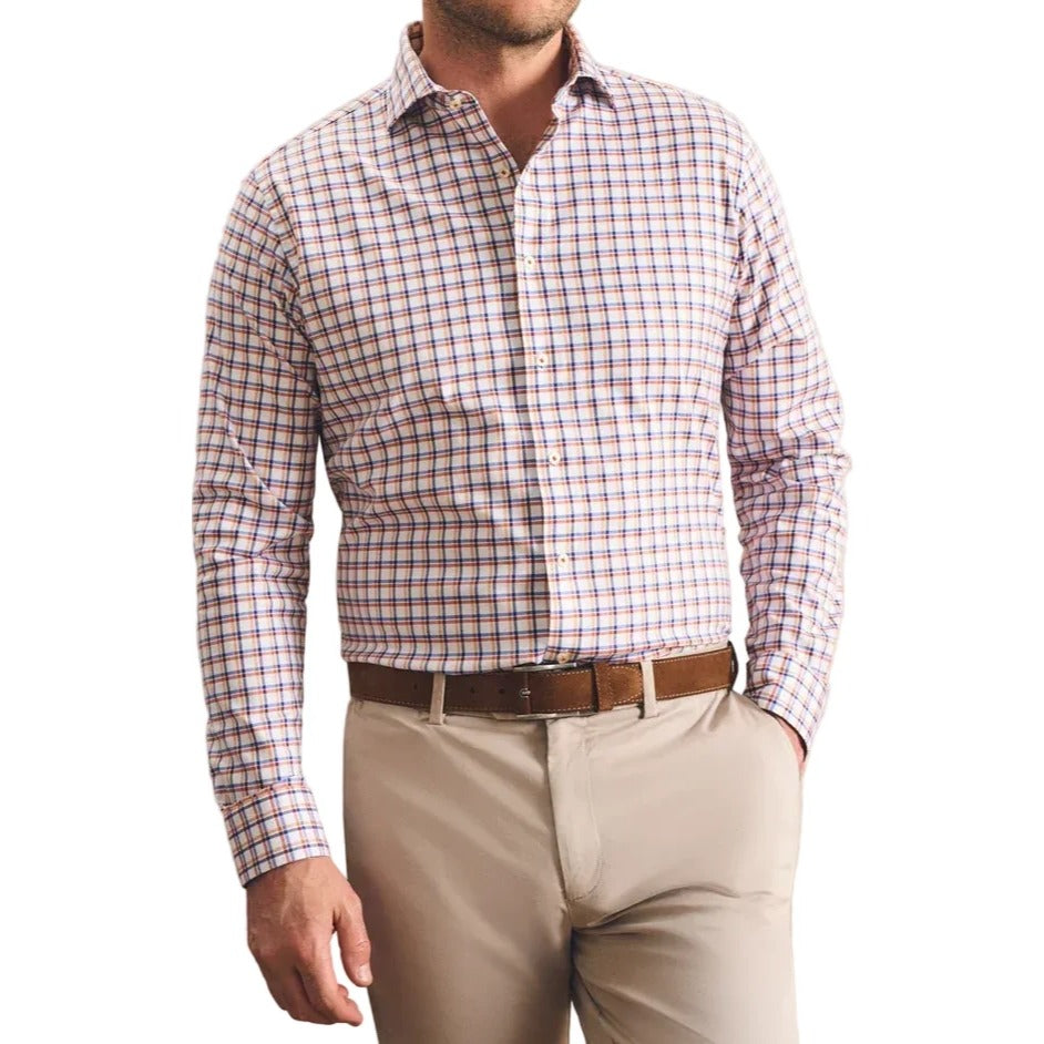A man wearing a long-sleeve button-down shirt with a red, blue, and white gingham pattern, paired with beige trousers and a brown belt, standing against a textured beige wall.