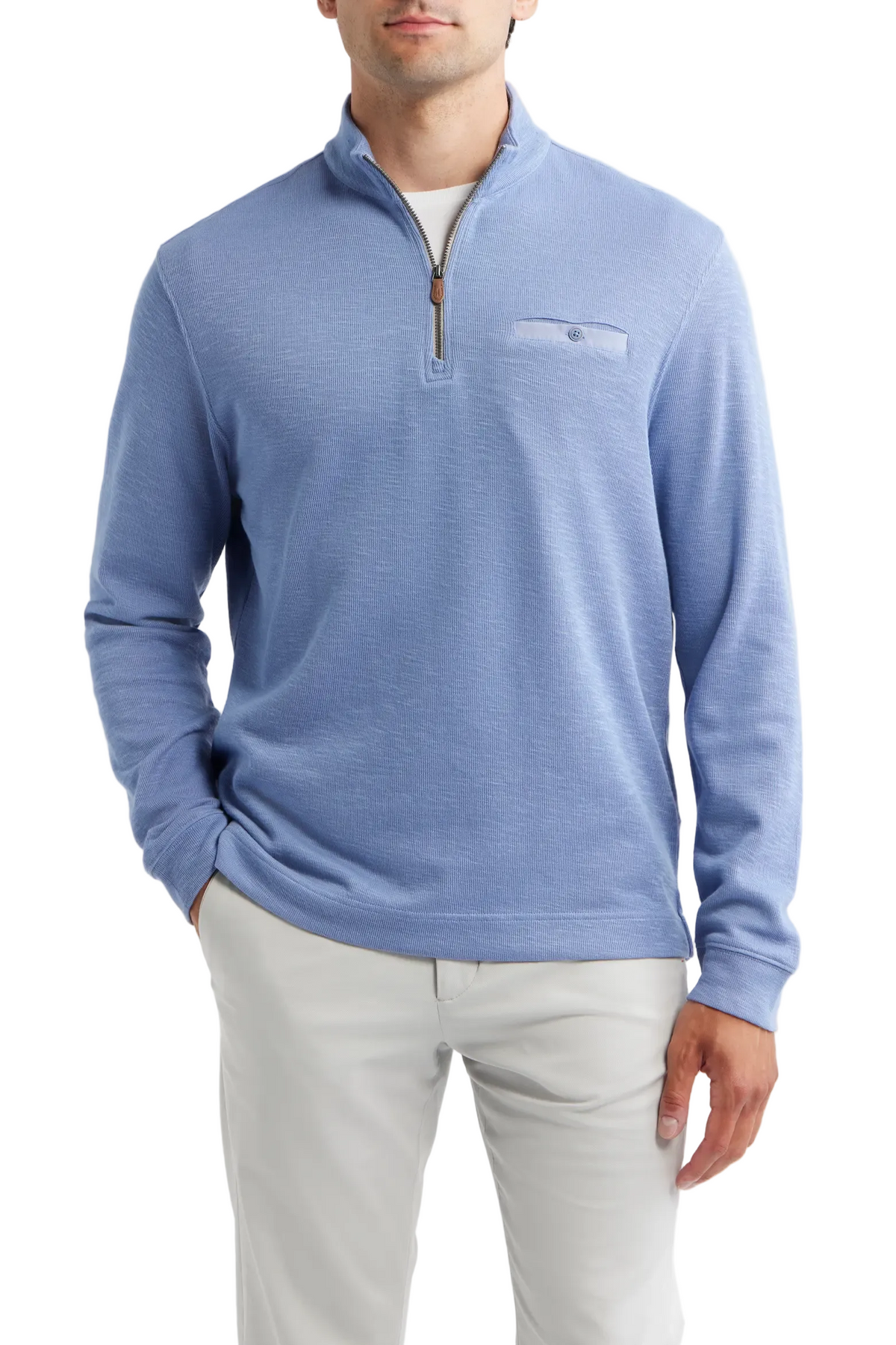 A man wearing a light blue quarter-zip knit sweater with a chest pocket, paired with light gray trousers, against a plain light gray background.