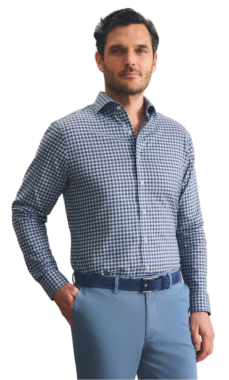 A man wearing a blue and white gingham long-sleeve dress shirt, paired with light blue trousers and a dark blue belt, standing against a textured beige wall.