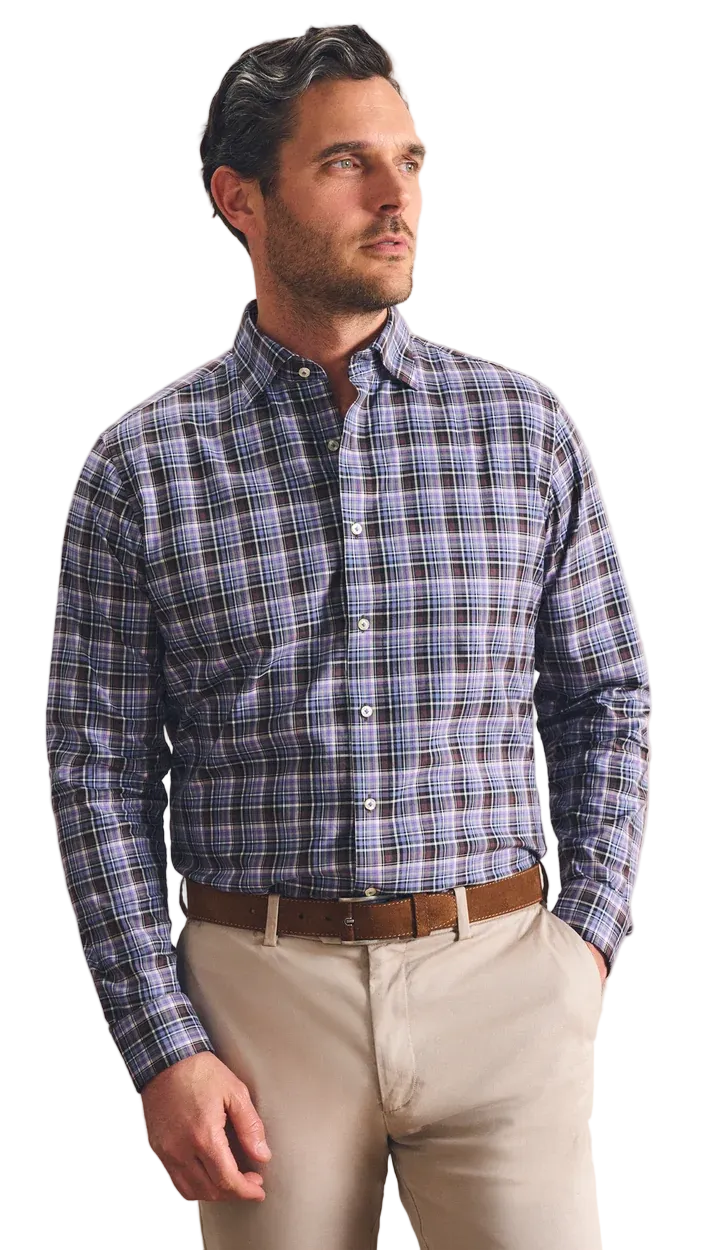 A man wearing a blue and gray plaid button-down shirt, beige trousers, and a brown belt, standing against a textured beige wall.