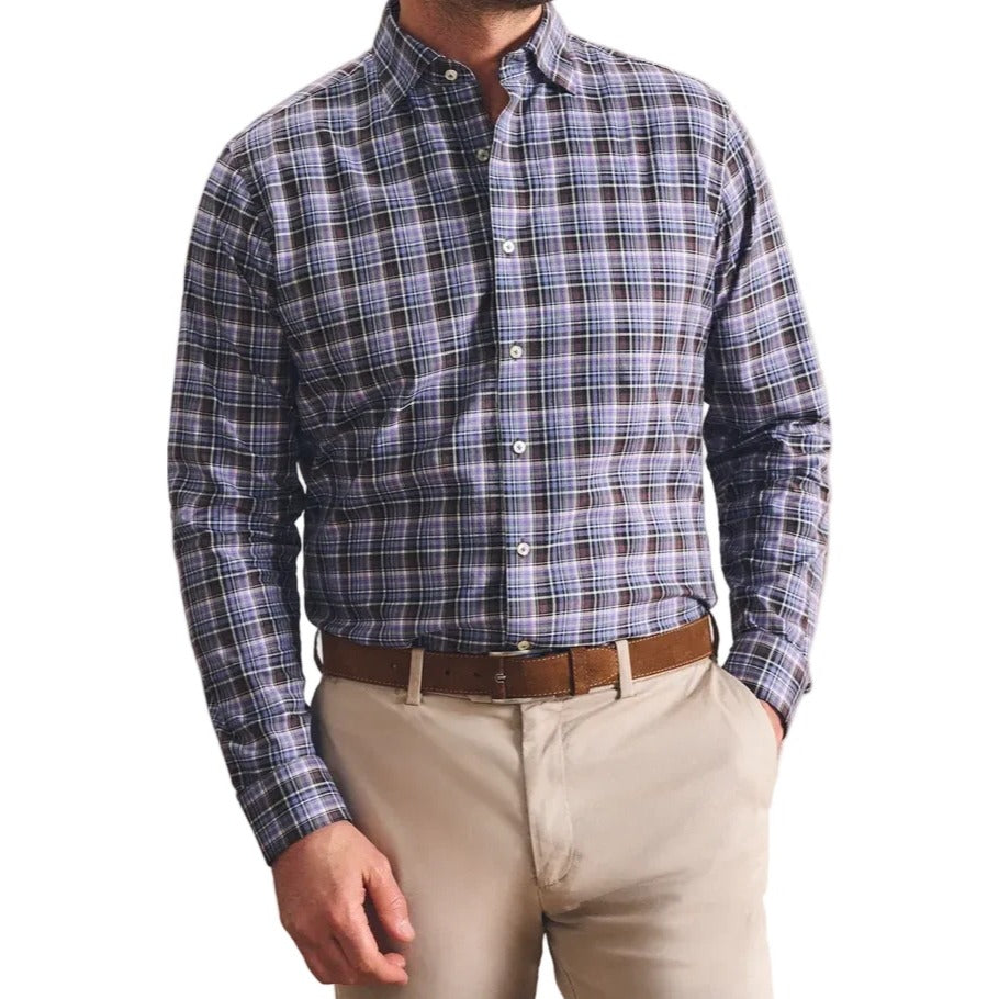 A man wearing a blue and gray plaid button-down shirt, beige trousers, and a brown belt, standing against a textured beige wall.