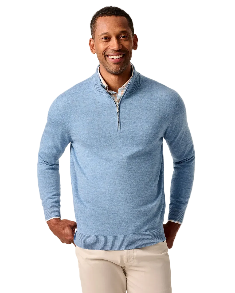 A smiling man wearing a light blue quarter-zip knit sweater over a white collared shirt and cream pants, standing against a plain light beige studio background.