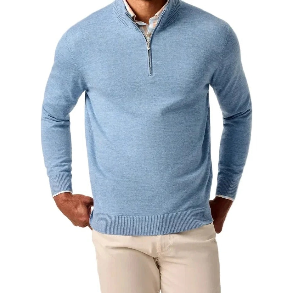 A smiling man wearing a light blue quarter-zip knit sweater over a white collared shirt and cream pants, standing against a plain light beige studio background.