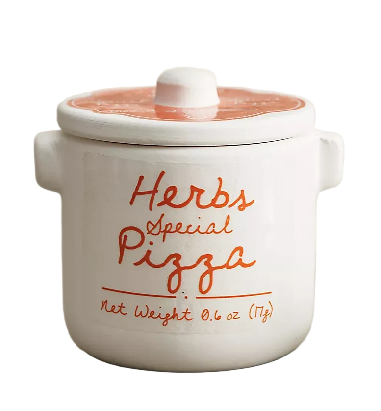 A white ceramic jar with a lid, featuring orange text that reads 'Herbs Special Pizza' and 'Net Weight 0.6 oz (17g)', set on a gray surface with blurred flowers and a bottle in the background.