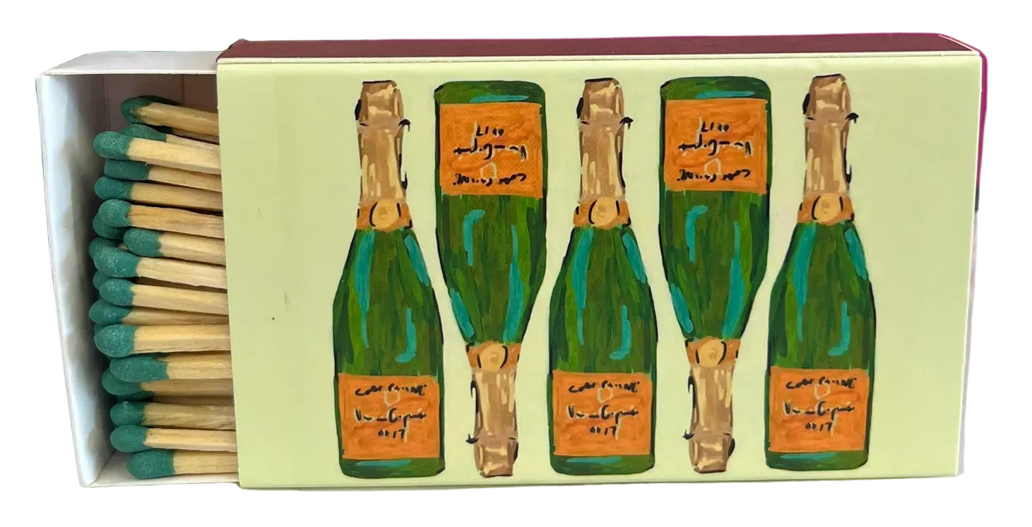 A decorative matchbox with illustrated green champagne bottles and Arabic script, shown open with matches on a marble surface against a bright pink wall and green plant.