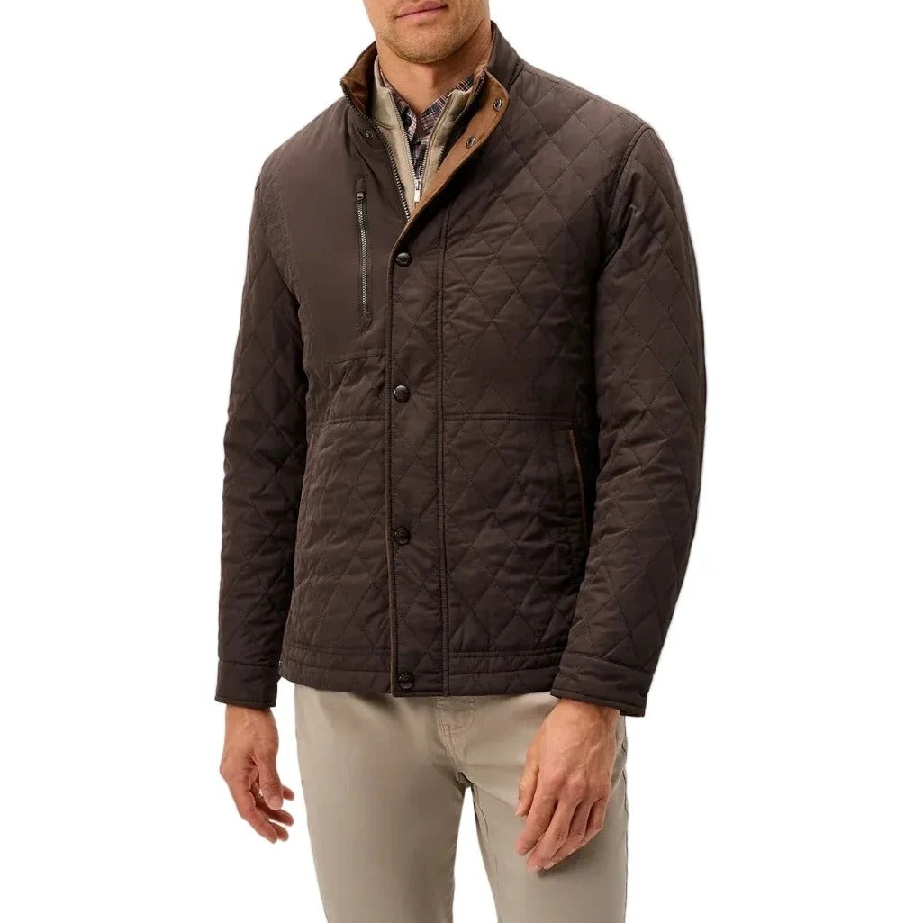 A man wearing a dark brown diamond-quilted jacket with a tan-lined collar over a patterned shirt and beige trousers, standing against a light gray studio background.
