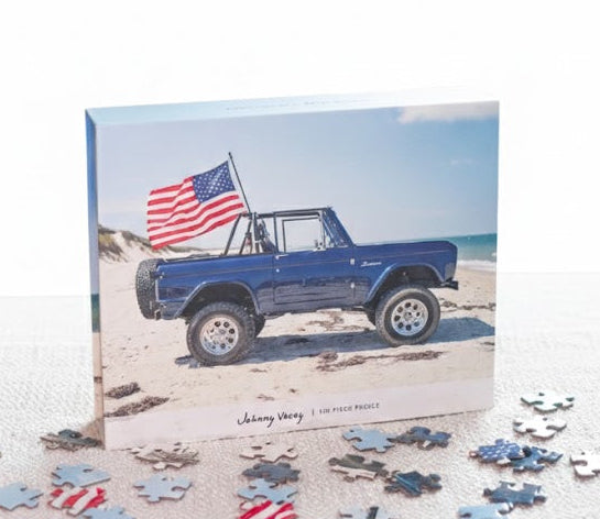 Puzzle box with a blue truck and American flag on a beach scene, surrounded by scattered puzzle pieces.
